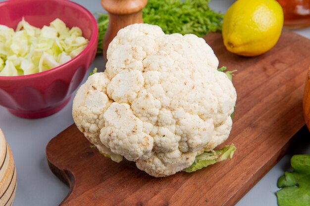 Vista cercana de coliflor y limón en la tabla de cortar con repollo en rodajas en un tazón montón de cilantro sobre fondo azul.