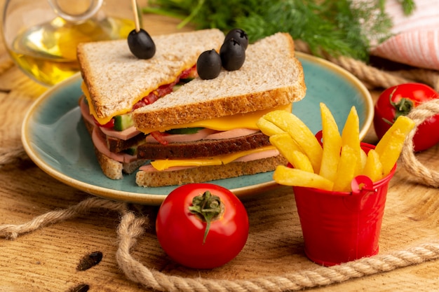 Vista cercana frontal sabroso sándwich con aceitunas, jamón y tomates dentro de la placa junto con papas fritas