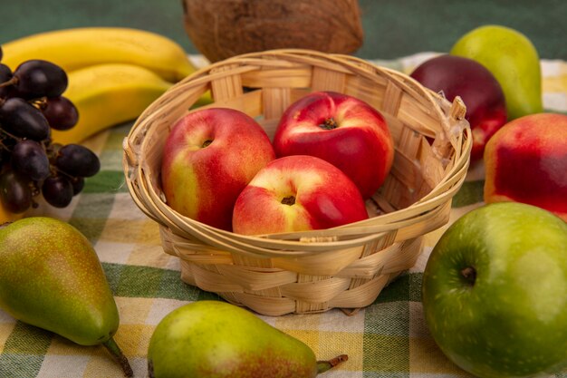 Vista cercana de frutas como melocotón en canasta y coco plátano pera uva en tela escocesa sobre fondo verde