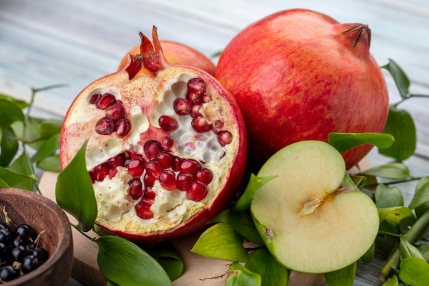 Vista cercana de frutas como mitades de granada y manzana con enteras y tazón de endrinas con hojas sobre superficie de madera