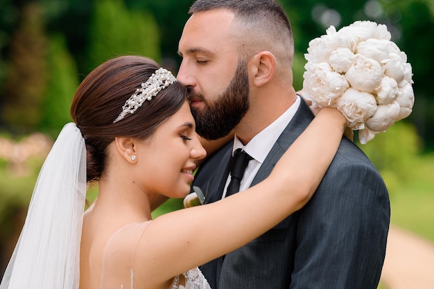 Vista cercana del guapo novio barbudo en traje cerrando los ojos y besando a la novia que sonríe y lo abraza en el cuello con un ramo de flores durante la caminata de la boda en la naturaleza