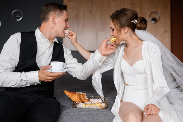 Vista cercana de una hermosa pareja con ropa de moda sentada en la cama cerca del plato con croissants y galletas alimentándose mutuamente con macarons y pasando la mañana de la boda juntos en la habitación del hotel