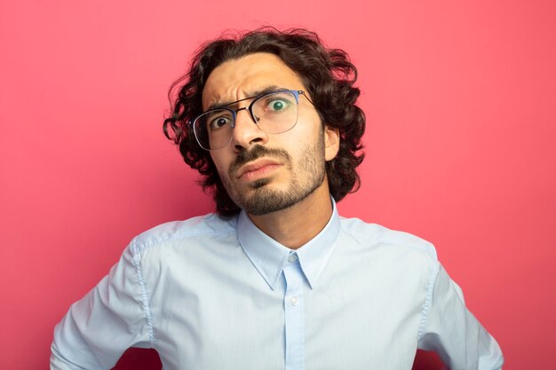 Vista cercana del joven apuesto joven impresionado con gafas mirando al frente aislado en la pared rosa