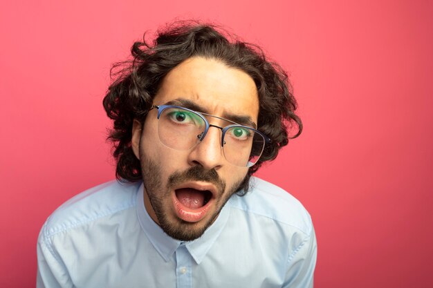 Vista cercana del joven apuesto joven impresionado con gafas mirando al frente aislado en la pared rosa