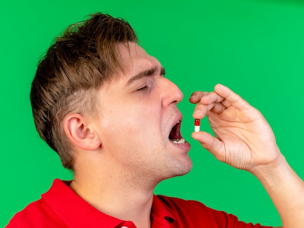 Vista cercana de joven guapo rubio enfermo tomando píldora médica con los ojos cerrados aislados en la pared verde