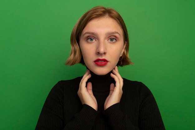 Vista cercana de la joven mujer rubia mirando al frente tocando el cuello aislado en la pared verde