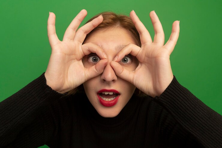 Foto gratuito vista cercana de la joven rubia impresionada mirando al frente haciendo gesto de mirada con las manos como binoculares aislados en la pared verde