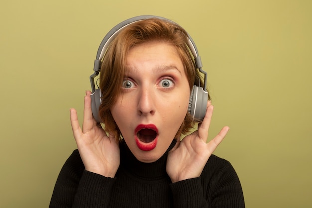 Vista cercana de la joven rubia sorprendida usando y tocando auriculares aislados en la pared verde oliva