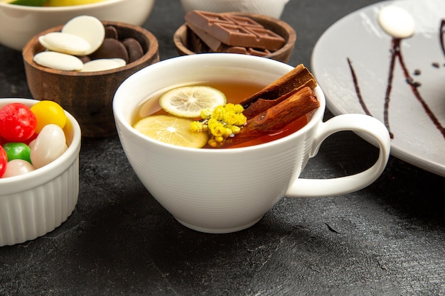 Vista cercana de lado una taza de té una taza de té con palitos de canela y limón junto a los cuencos de caramelos en la mesa oscura