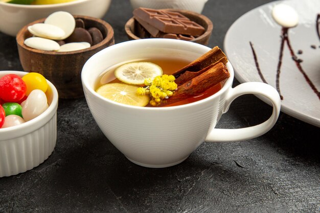 Vista cercana de lado una taza de té una taza de té con palitos de canela y limón junto a los cuencos de caramelos en la mesa oscura