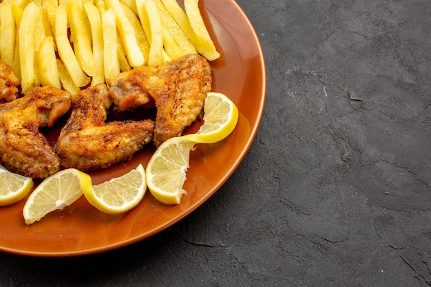 Vista cercana lateral fastfood naranja plato de un apetitoso alitas de pollo papas fritas y limón sobre el fondo oscuro