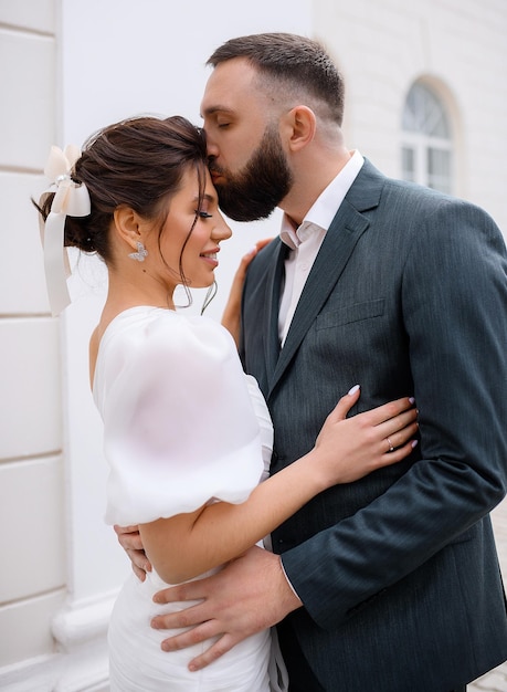 Vista cercana de una linda chica con un vestido blanco de moda con el pelo atado y un lazo en la cabeza parada cerca de su marido y cerrando los ojos mientras él besa su cara y la abraza al aire libre