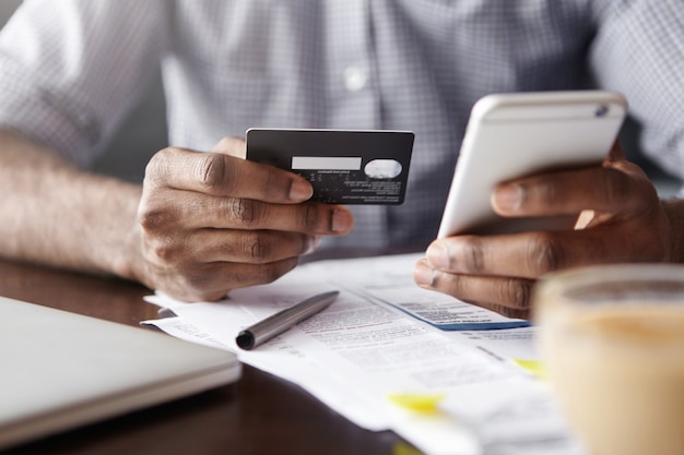 Vista cercana de las manos del hombre africano con tarjeta de crédito de plástico y smartphone