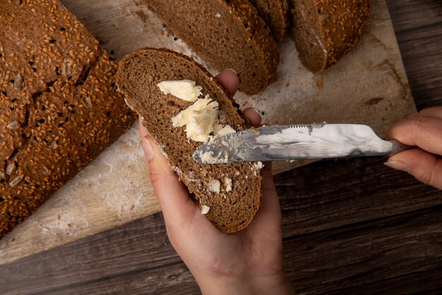 Vista cercana de manos de mujer untando mantequilla en una rebanada de pan con un cuchillo y cortar el pan en la superficie de madera y fondo