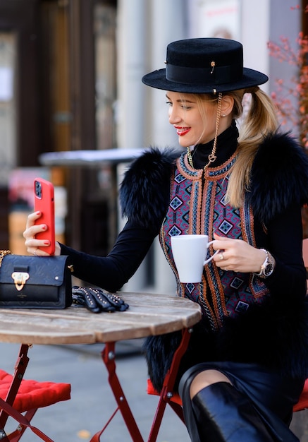 Vista cercana de una mujer atractiva con sombrero negro con cadena vestida con chaleco de moda con piel y bordado sentado en la mesa usando el teléfono móvil y tomando selfie mientras bebe café en la cafetería