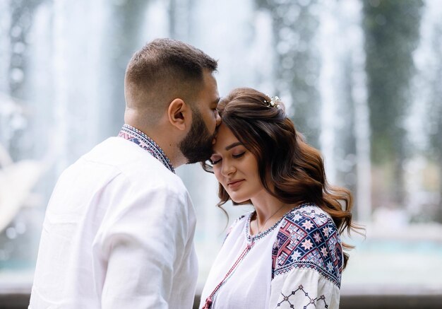 Vista cercana de una mujer deslumbrante y pensativa con maquillaje natural y cabello rizado vestido con una camisa con un patrón colorido inclinando la cabeza hacia adelante mientras el hombre besaba su rostro en el fondo de las fuentes