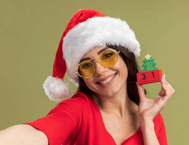 Vista cercana de la niña bonita joven sonriente con gorro de Papá Noel y gafas sosteniendo el juguete del árbol de Navidad con la fecha estirando la mano aislada en la pared verde oliva