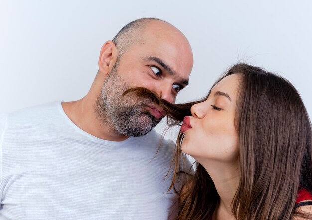 Vista cercana de la pareja adulta hombre juguetón haciendo bigote de cabello de mujer mirando a su mujer haciendo gesto de beso