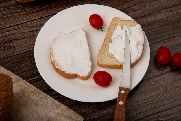 Vista cercana de la placa de rebanadas de pan blanco con queso cottage y tomates y cuchillo sobre fondo de madera