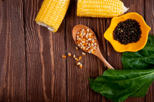 Vista cercana de semillas de maíz en cuchara y semillas de pimienta negra en un tazón con callos y espinacas en la mesa de madera con espacio de copia