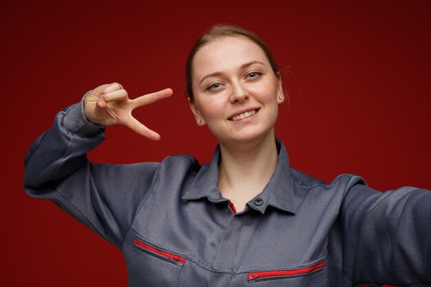 Vista cercana de la sonriente joven ingeniera rubia vistiendo uniforme extendiendo la mano hacia la cámara mostrando el símbolo de signo v cerca del ojo