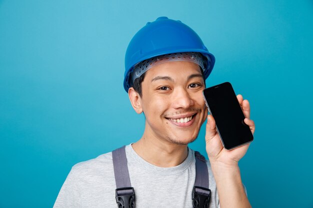 Vista cercana de sonriente joven trabajador de la construcción con casco de seguridad y uniforme sosteniendo teléfono móvil