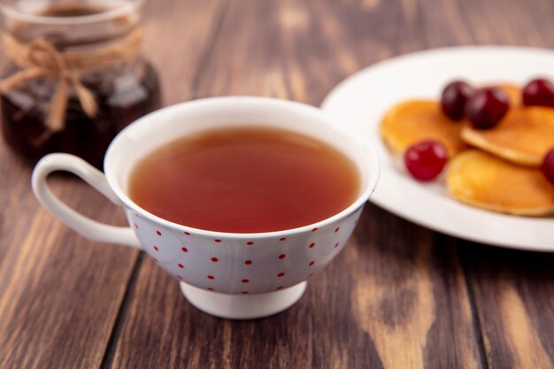 Vista cercana de la taza de té con plato de panqueques y tarro de mermelada de fresa sobre fondo de madera