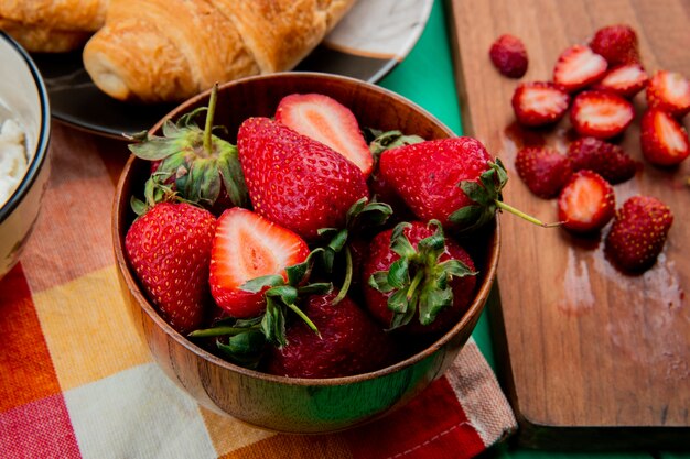 Vista cercana del tazón de fresas con media luna roll en placa sobre tela y cortar fresas en tabla de cortar