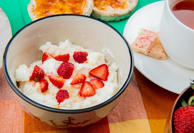 Vista cercana del tazón de requesón con fresas taza de té pan crujiente sobre tela