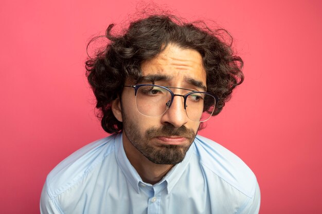 Vista cercana de triste joven guapo con gafas mirando al lado aislado en la pared rosa