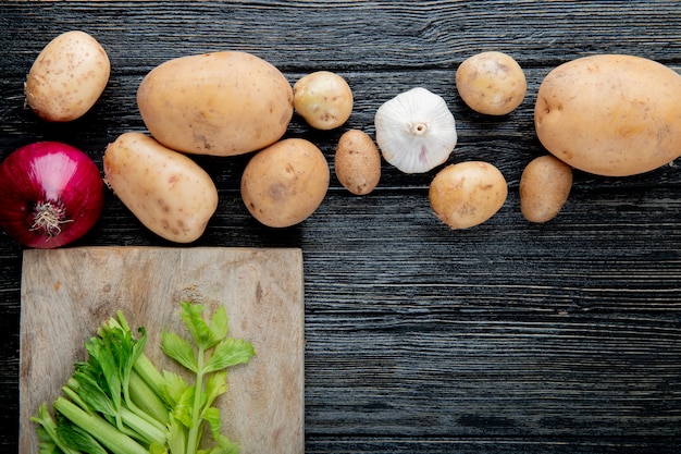 Vista cercana de verduras como cebolla patata ajo con apio cortado en la tabla de cortar sobre fondo de madera con espacio de copia