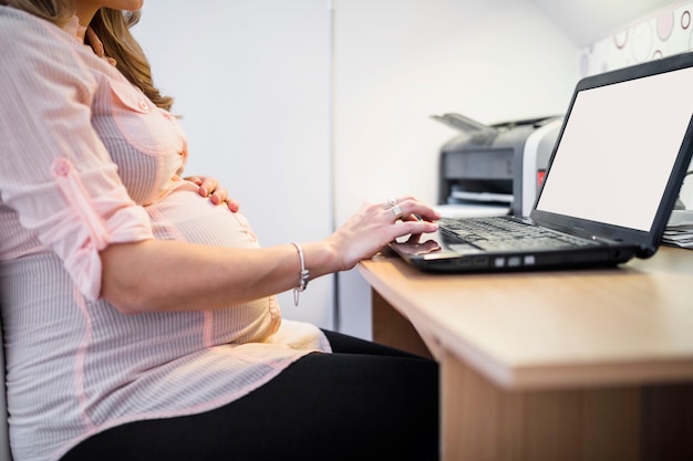 Vista de cintura de una mujer embarazada usando la computadora portátil en el escritorio de madera