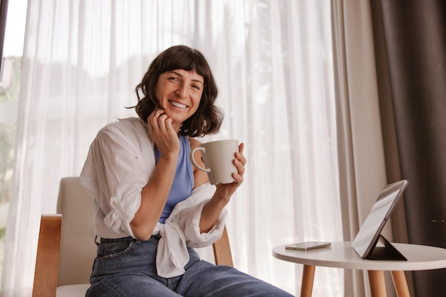 Vista completa de la mujer sonriente mirando a la cámara y sosteniendo una taza blanca