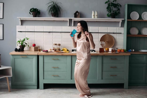 Vista desde el costado de una joven en pijama parada en la cocina y bailando por la mañana Mujer alegre cantando mientras prepara el desayuno en casa los fines de semana Concepto de preparación y felicidad