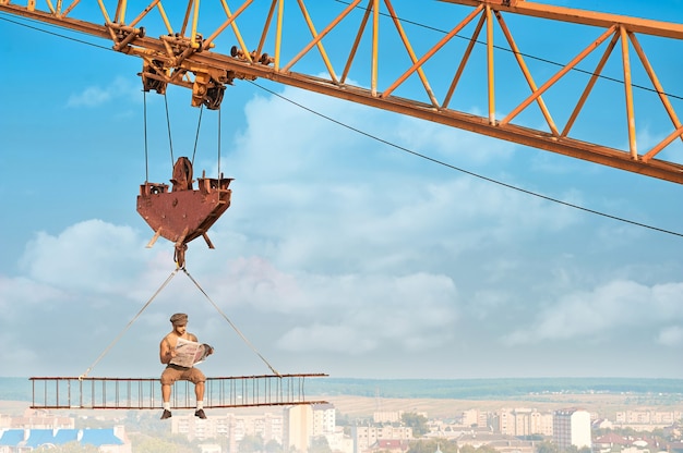 Vista desde la distancia del audaz constructor en ropa de trabajo sentado en la construcción en alto y leyendo el periódico. Grúa de construcción con construcción con hombre sobre la ciudad. Construcción extrema de casa en la gran ciudad.