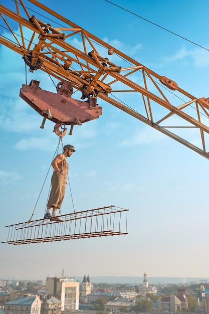 Vista desde la distancia del constructor atlético en ropa de trabajo de pie en la construcción en lo alto. Grúa de construcción con construcción con hombre sobre la ciudad. Construcción extrema de casa en la gran ciudad. Hombre mirando hacia abajo.