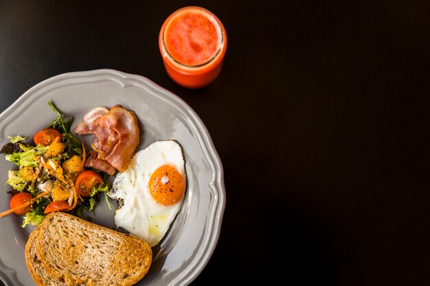 Una vista elevada de batido rojo en frasco de vidrio con pan tostado; ensalada; tocino y huevo frito en placa gris sobre fondo negro