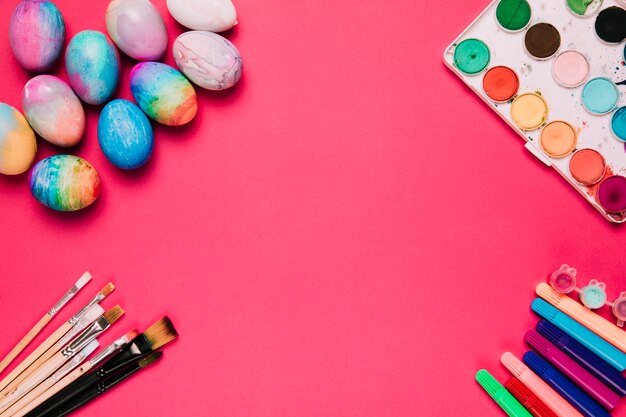 Una vista elevada de coloridos huevos de pascua; cepillos rotulador y caja de pintura de acuarela sobre fondo rosa