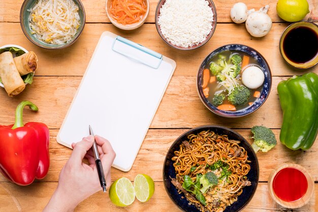 Una vista elevada de la comida tailandesa con una persona que escribe en el portapapeles con pluma