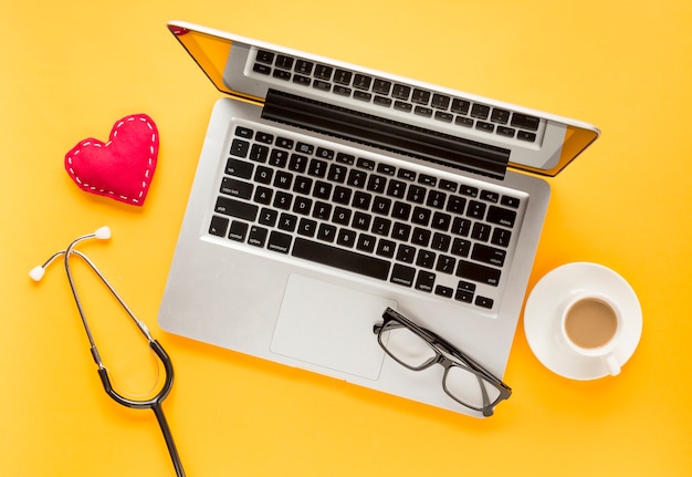 Vista elevada de la computadora portátil con anteojos; corazón cosido Taza de té y estetoscopio sobre fondo amarillo