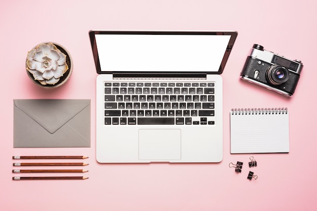 Vista elevada de la computadora portátil; cámara; Papelerías y planta suculenta sobre fondo rosa