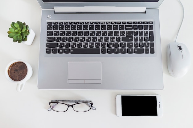 Vista elevada de la computadora portátil con el ratón, la taza de café, las lentes y el teléfono móvil sobre el escritorio blanco