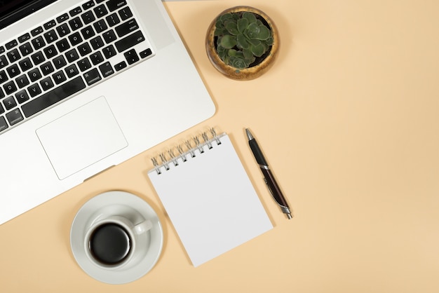 Vista elevada de la computadora portátil; taza de café; bolígrafo; y la libreta espiral sobre fondo beige