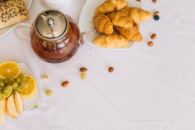 Una vista elevada de croissant al horno; frutas Té y frutos secos sobre mantel blanco
