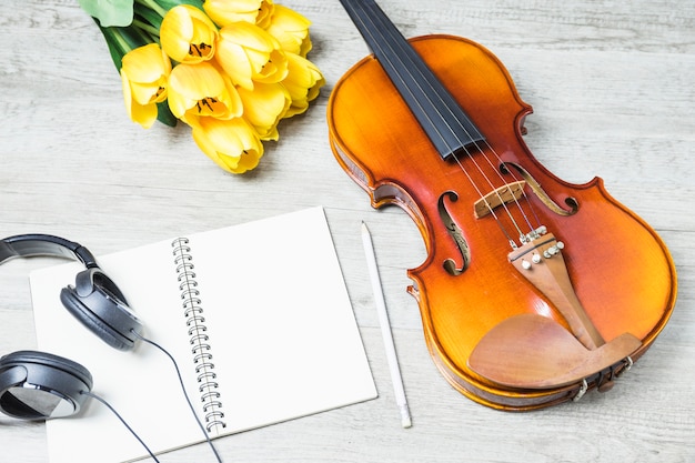Vista elevada del cuaderno en blanco con auriculares; lápiz; tulipán y violín clásico en el fondo de madera