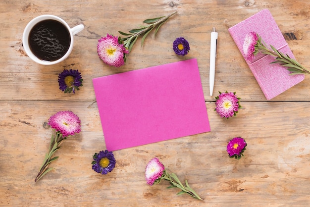 Vista elevada de hermosas flores; papel rosa en blanco; bolígrafo; Diario y té negro sobre superficie de madera.