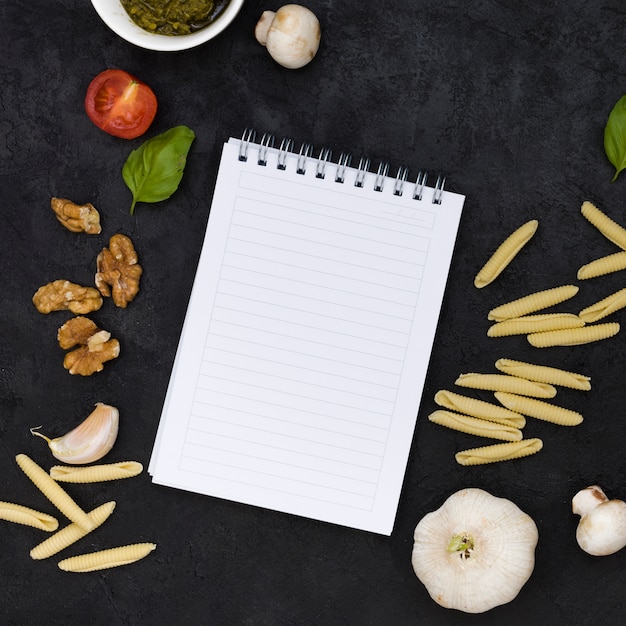 Una vista elevada de la libreta espiral en blanco con pasta garganelli; nueces albahaca; tomate; Champiñones y dientes de ajo sobre fondo negro