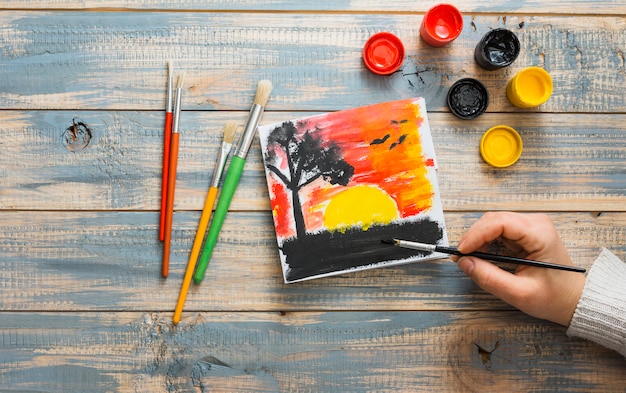 Vista elevada de la mano humana pintando el atardecer visto con pincel en el escritorio de madera