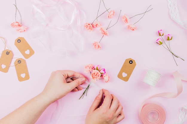 Una vista elevada de la mano humana que ata las flores artificiales con cinta sobre fondo rosa