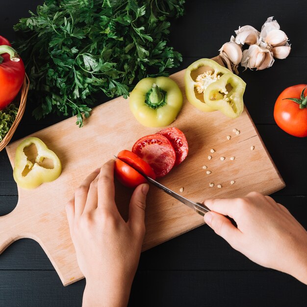 Vista elevada de una mano humana que corta un tomate rojo fresco en una tabla de cortar de madera